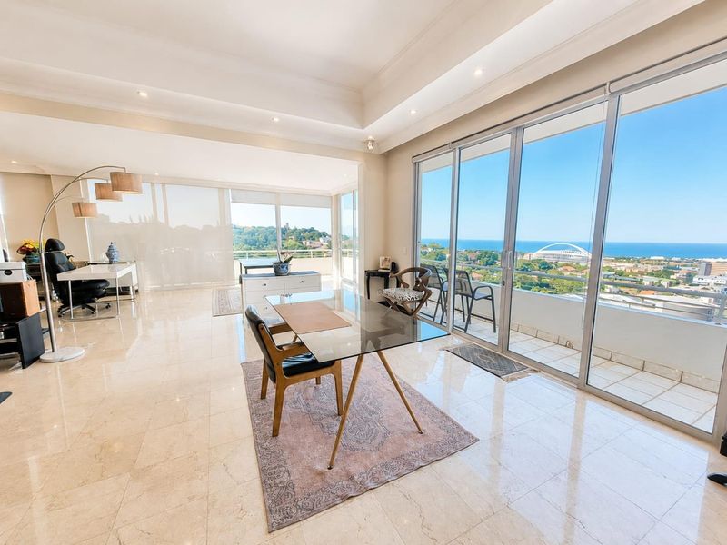 Spacious open-plan living area featuring large format floor tiles, tray ceilings with recessed lighting, and expansive sliding glass doors leading to a balcony with ocean views.