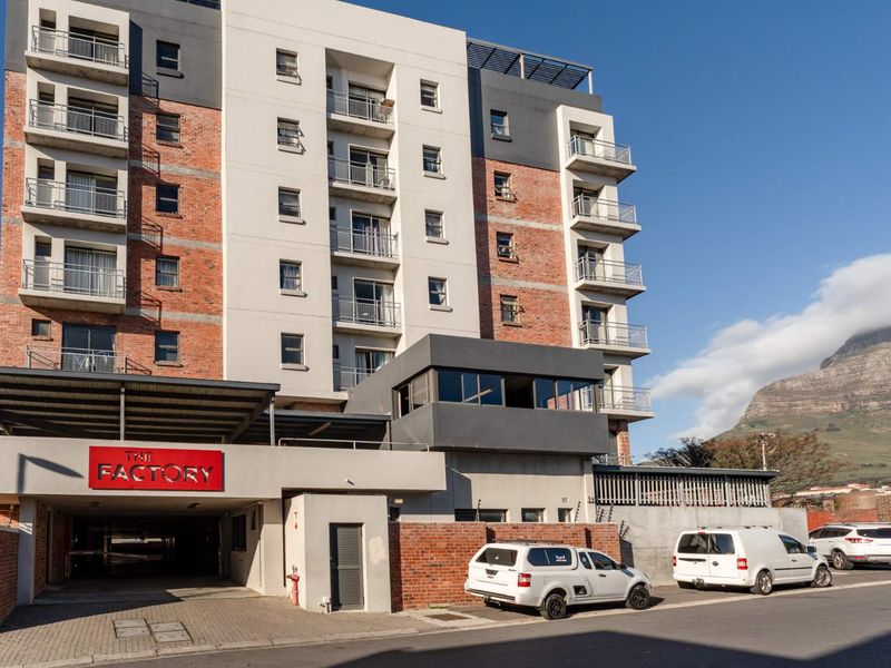 Exterior view of The Factory apartment building featuring a modern brick and concrete facade with multiple balconies and a covered parking entrance.