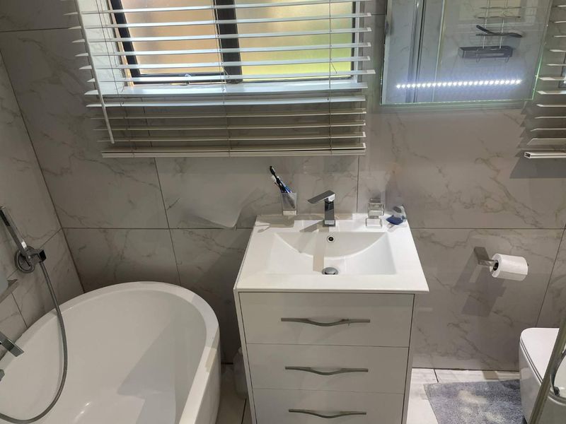 Modern bathroom featuring a sleek vanity cabinet, basin with mixer tap, freestanding bathtub with handheld shower, and illuminated mirror with marble-look wall tiles.