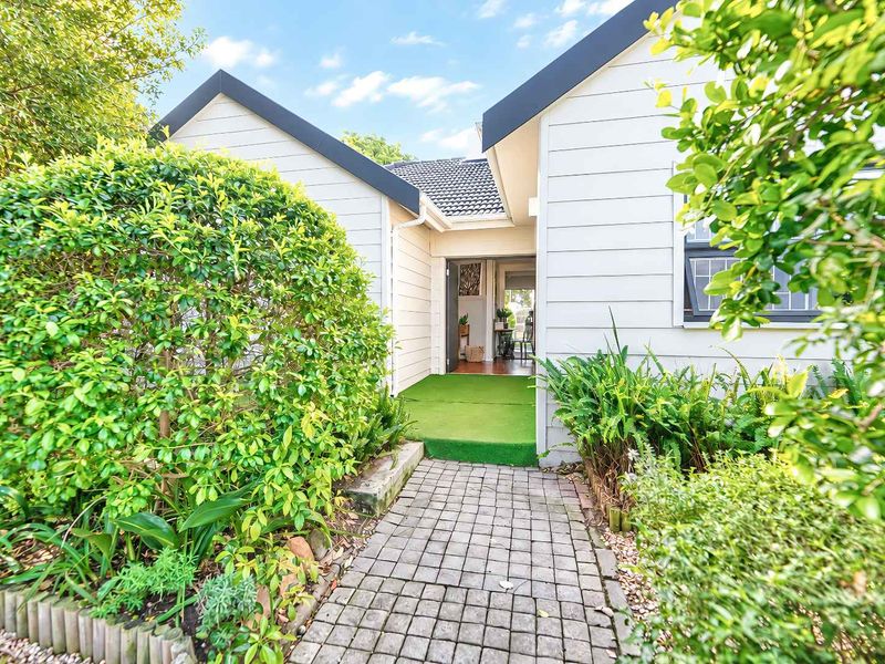 A welcoming front entrance featuring a paved walkway leading to the main door, framed by well-maintained timber cladding and established garden landscaping.