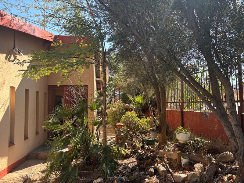 A rear garden area featuring mature trees, established landscaping, a paved walkway, and metal perimeter fencing along the property boundary.