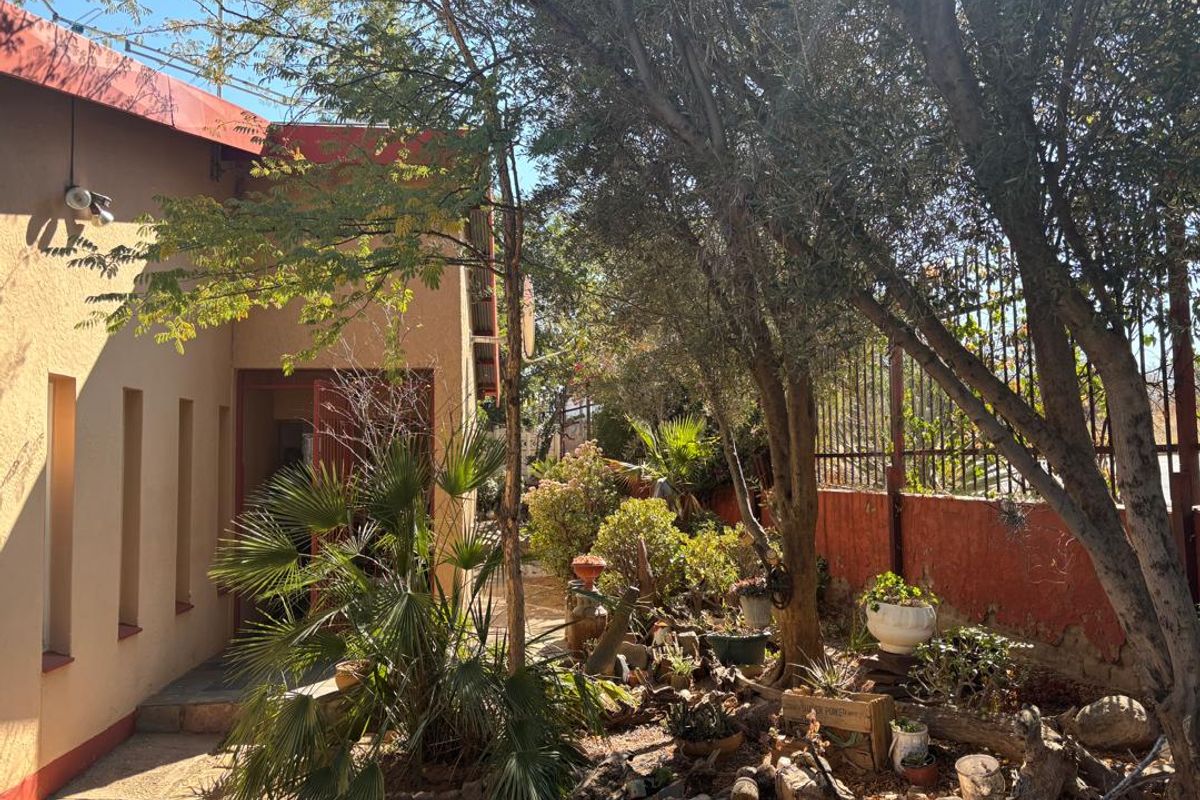 A rear garden area featuring mature trees, established landscaping, a paved walkway, and metal perimeter fencing along the property boundary.