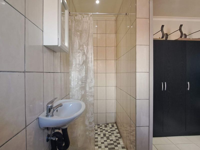 A functional bathroom featuring a wall-mounted basin, a shower area with a curtain, and tiled finishes, adjacent to a built-in storage cupboard.