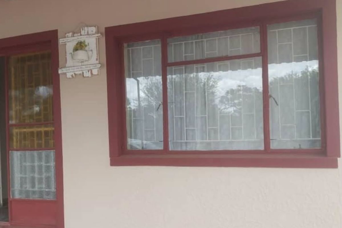 A covered patio area featuring a concrete floor, security gate, and window with burglar bars, showing signs of wear on the exterior finishes.