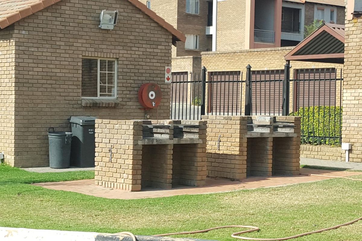 Communal outdoor area featuring built-in brick braai stands, paved flooring, and security fencing within the apartment complex grounds.