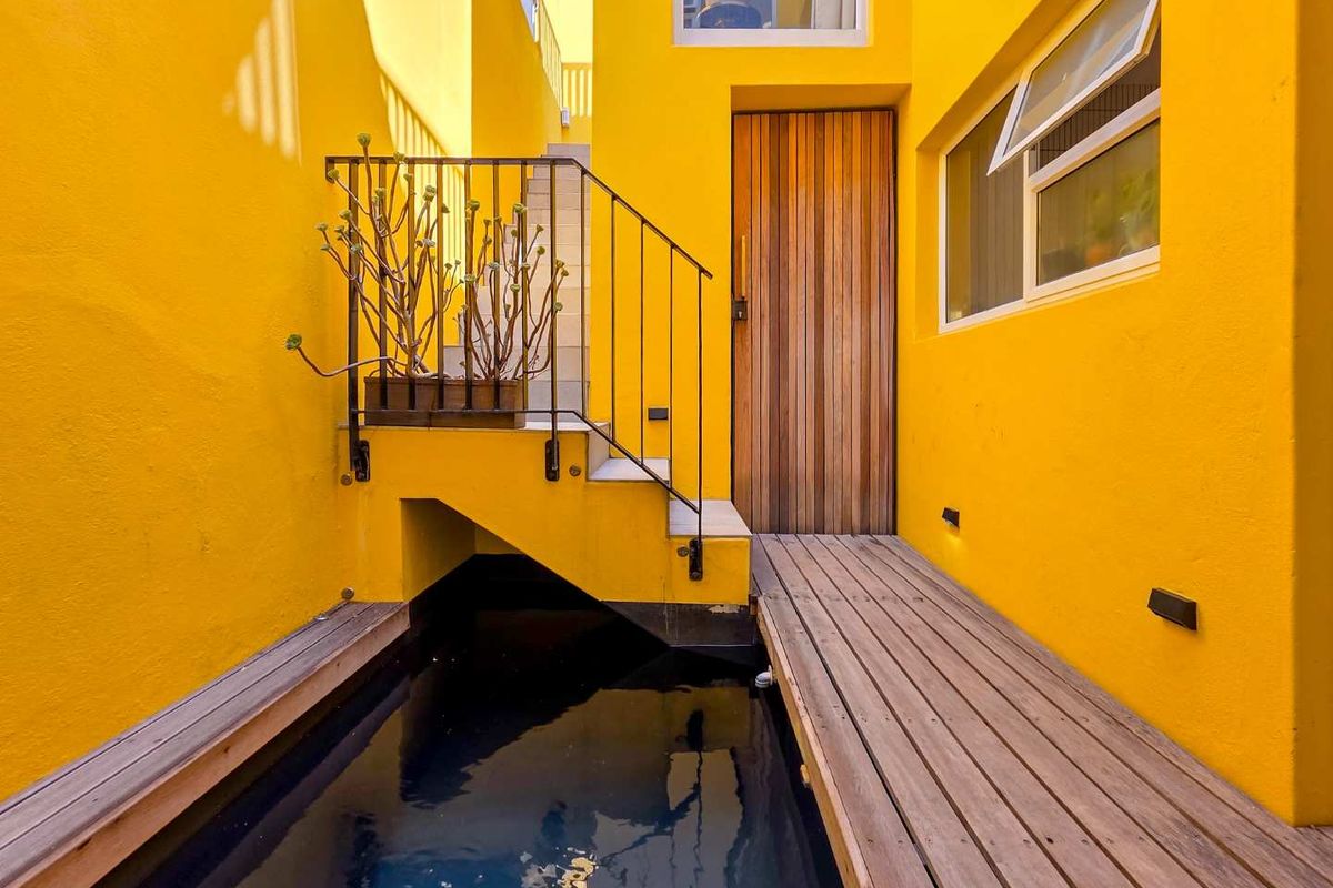 A vibrant yellow exterior courtyard featuring a timber deck walkway, a small water feature, and a modern timber entrance door.