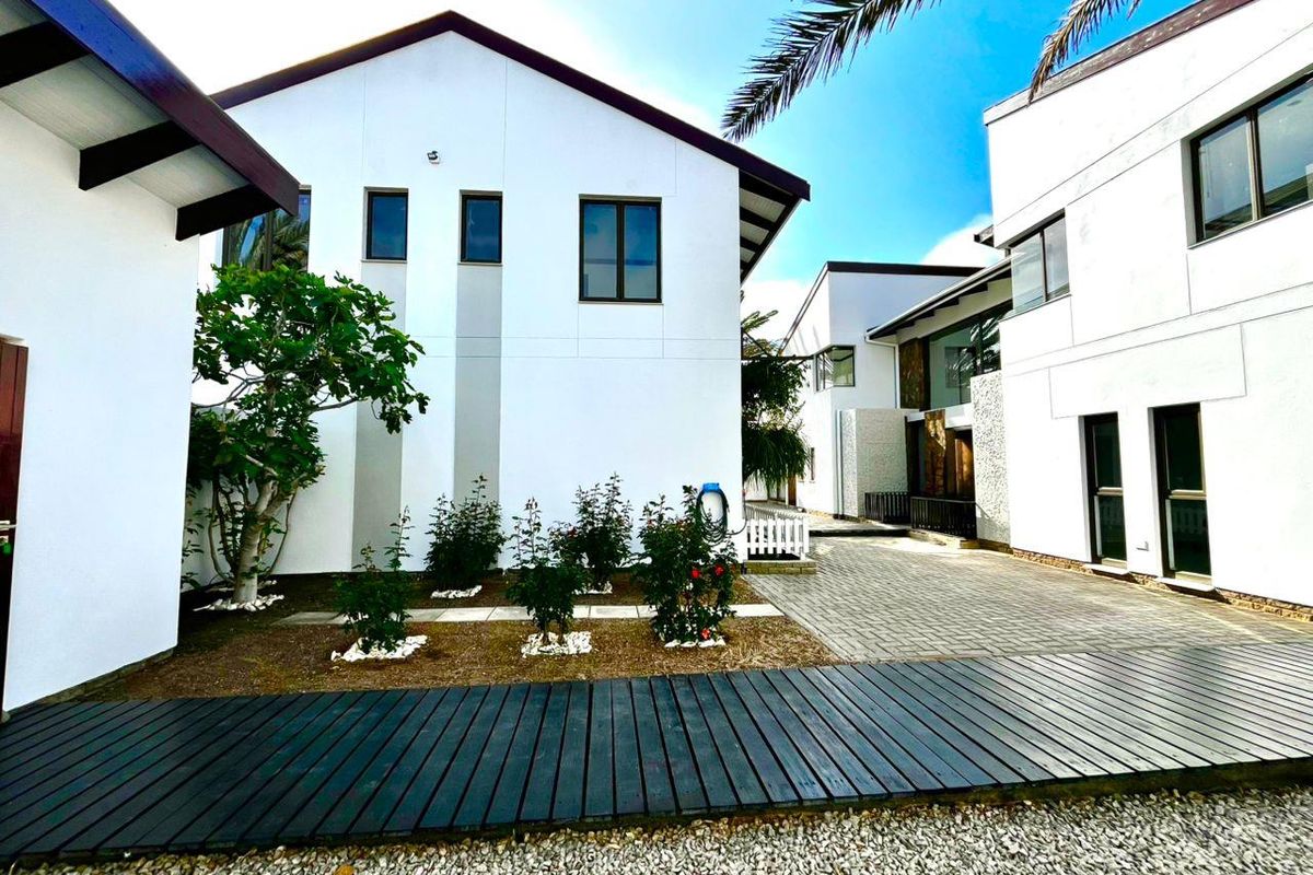 A modern exterior view of the property featuring white facades, dark window frames, a paved courtyard, and a wooden deck walkway.