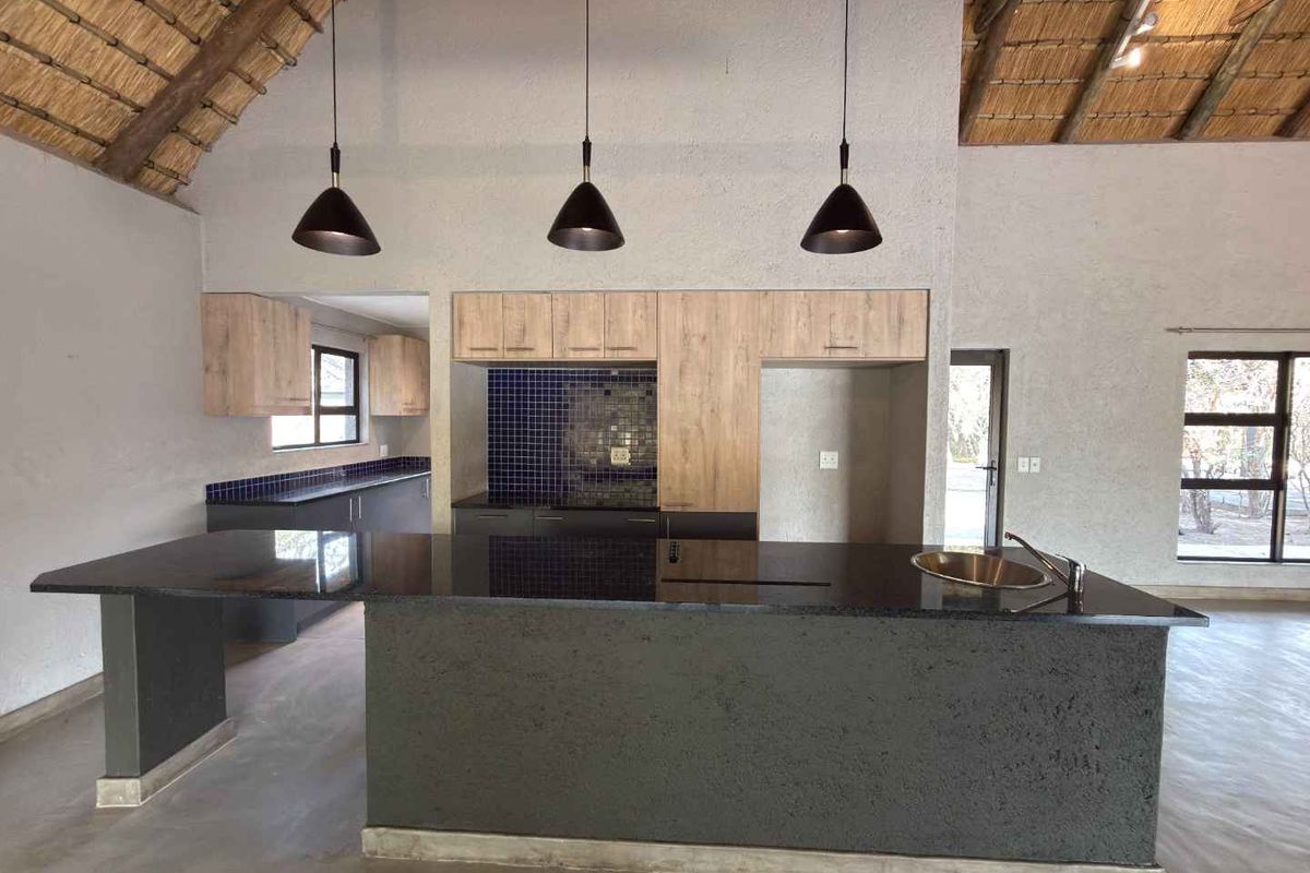 Modern open-plan kitchen featuring a large central island with a sink, dark stone countertops, wood-finish cabinetry, and a high-vaulted thatched ceiling.