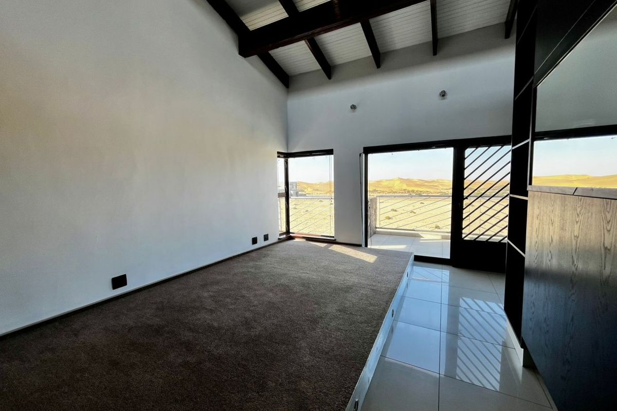 Spacious bedroom featuring a raised carpeted platform, exposed timber ceiling beams, built-in shelving, and sliding door access to a balcony with desert views.