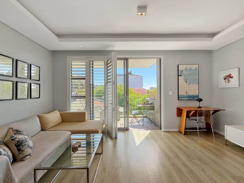 A bright and modern living room featuring laminate flooring, recessed ceiling lighting, and large sliding glass doors that open onto a private balcony.