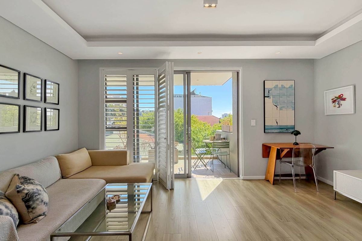 A bright and modern living room featuring laminate flooring, recessed ceiling lighting, and large sliding glass doors that open onto a private balcony.
