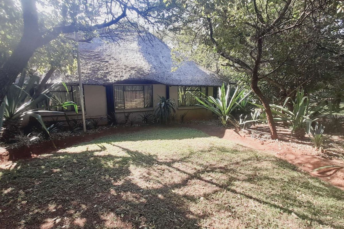 A view of the exterior rear facade featuring a traditional thatched roof structure surrounded by mature trees and landscaped garden beds.
