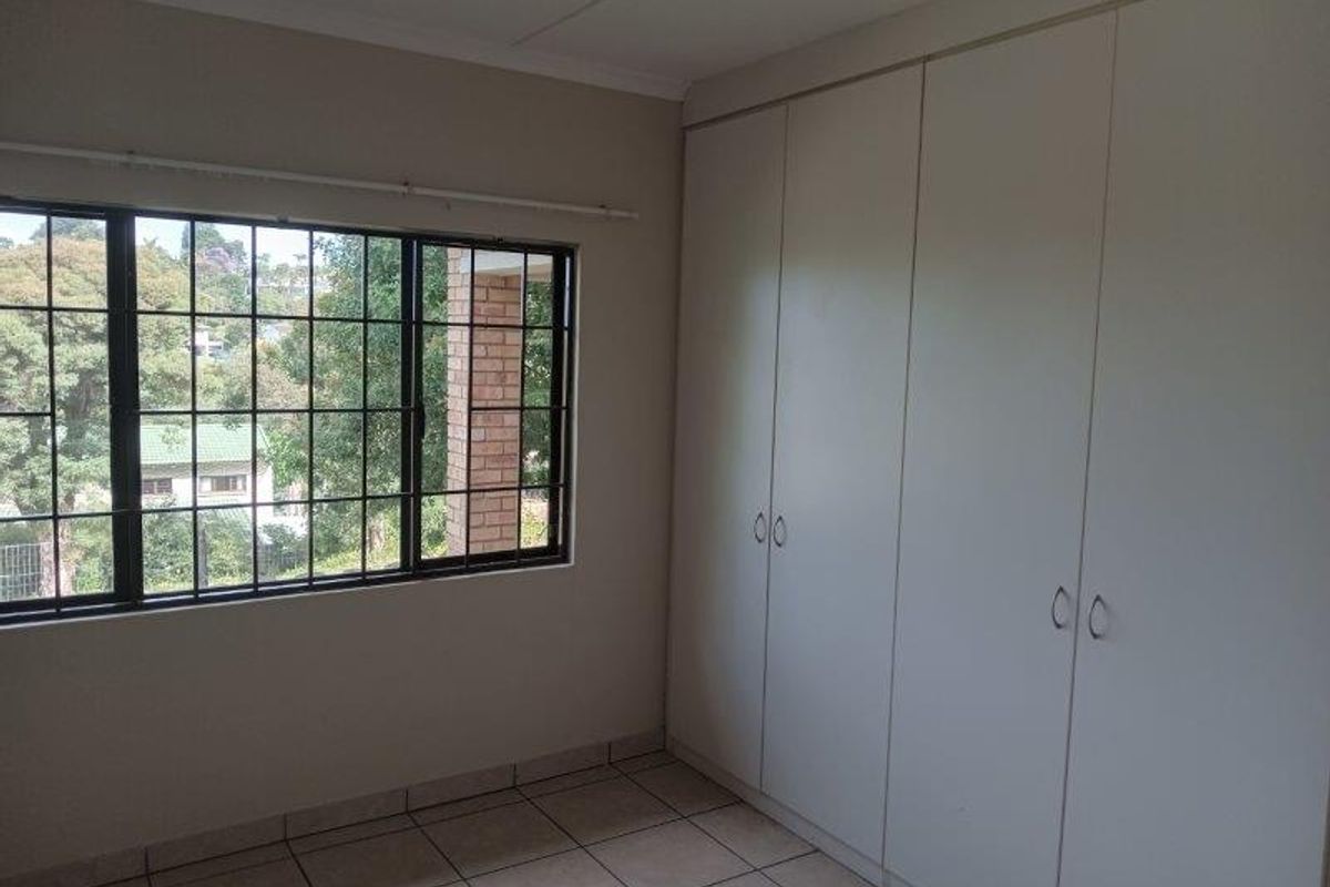 A bright, empty bedroom featuring neutral tiled flooring, large built-in cupboards, and a window with security bars overlooking the surrounding area.