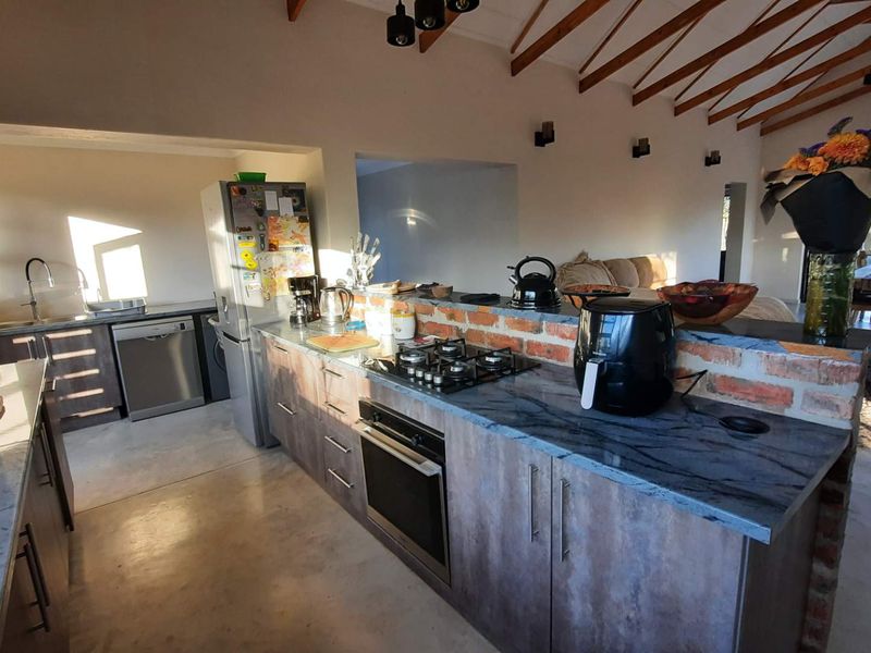 Spacious open-plan kitchen and living area featuring exposed timber rafters, concrete flooring, stone countertops, and a rustic exposed brick breakfast bar.