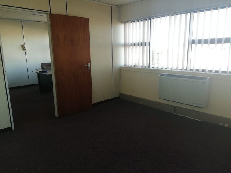 An empty office space featuring carpet flooring, vertical blinds, and a wall-mounted HVAC unit, with an adjacent office room visible through the doorway.