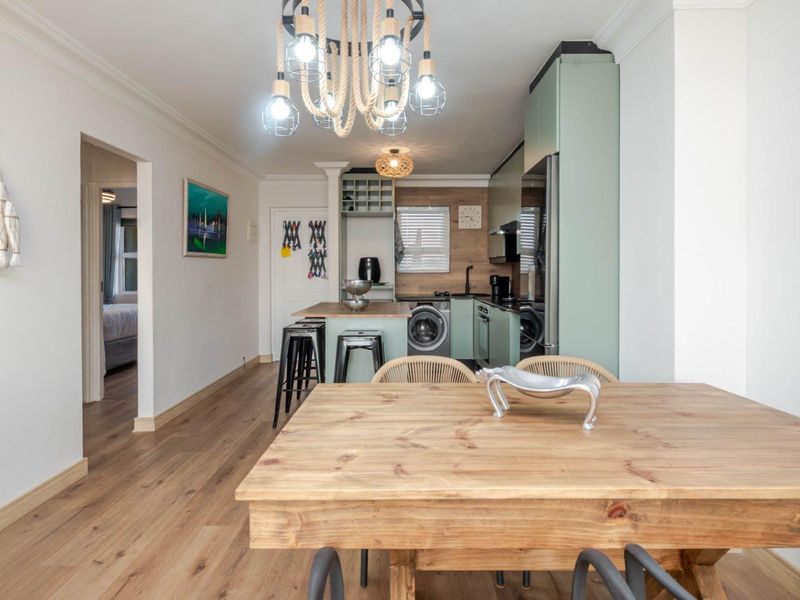 Modern open-plan kitchen and dining area featuring light green cabinetry, wood-look laminate flooring, and a functional kitchen island with integrated laundry appliances.