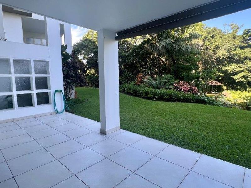 A spacious covered patio featuring light-colored floor tiles, a support pillar, and a retractable awning, overlooking a lush, well-maintained private garden.