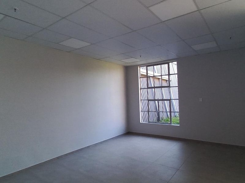 A spacious, empty office area featuring a suspended ceiling, tiled flooring, and a large window providing natural light.
