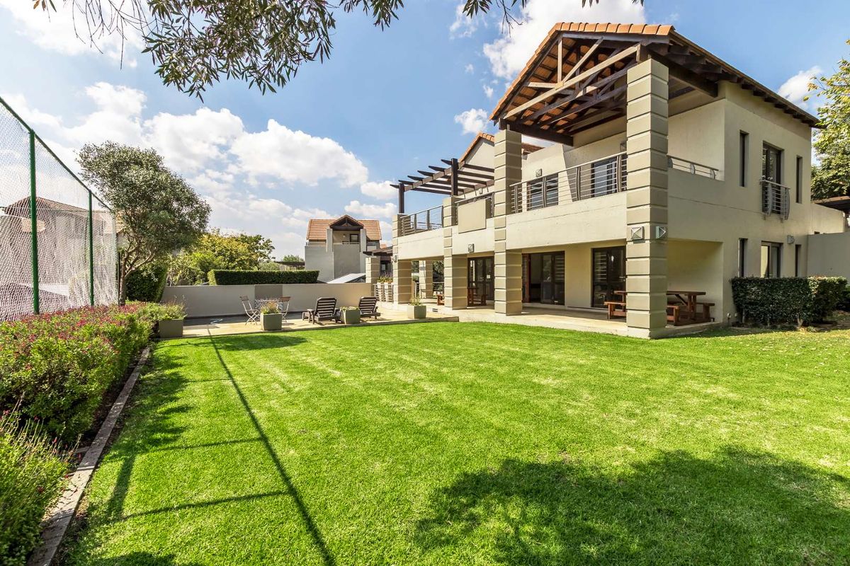 A spacious rear garden featuring a well-maintained lawn, paved patio area, and a two-story building with covered balconies and architectural pillars.