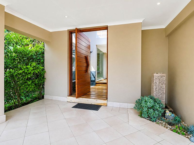 A welcoming covered entrance featuring a modern timber pivot door, tiled flooring, and a decorative stone water feature.