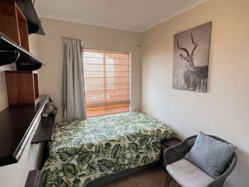 A compact bedroom featuring laminate flooring, built-in wall shelving, and a window with fitted blinds.