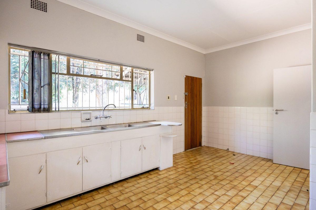 An empty, dated kitchen featuring original cabinetry, a stainless steel double sink, tiled splashbacks, and tiled flooring with security bars on the window.