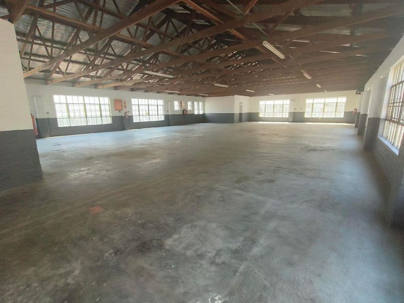 Spacious open-plan industrial warehouse area featuring exposed timber roof trusses, concrete flooring, and ample natural light through large steel-framed windows.