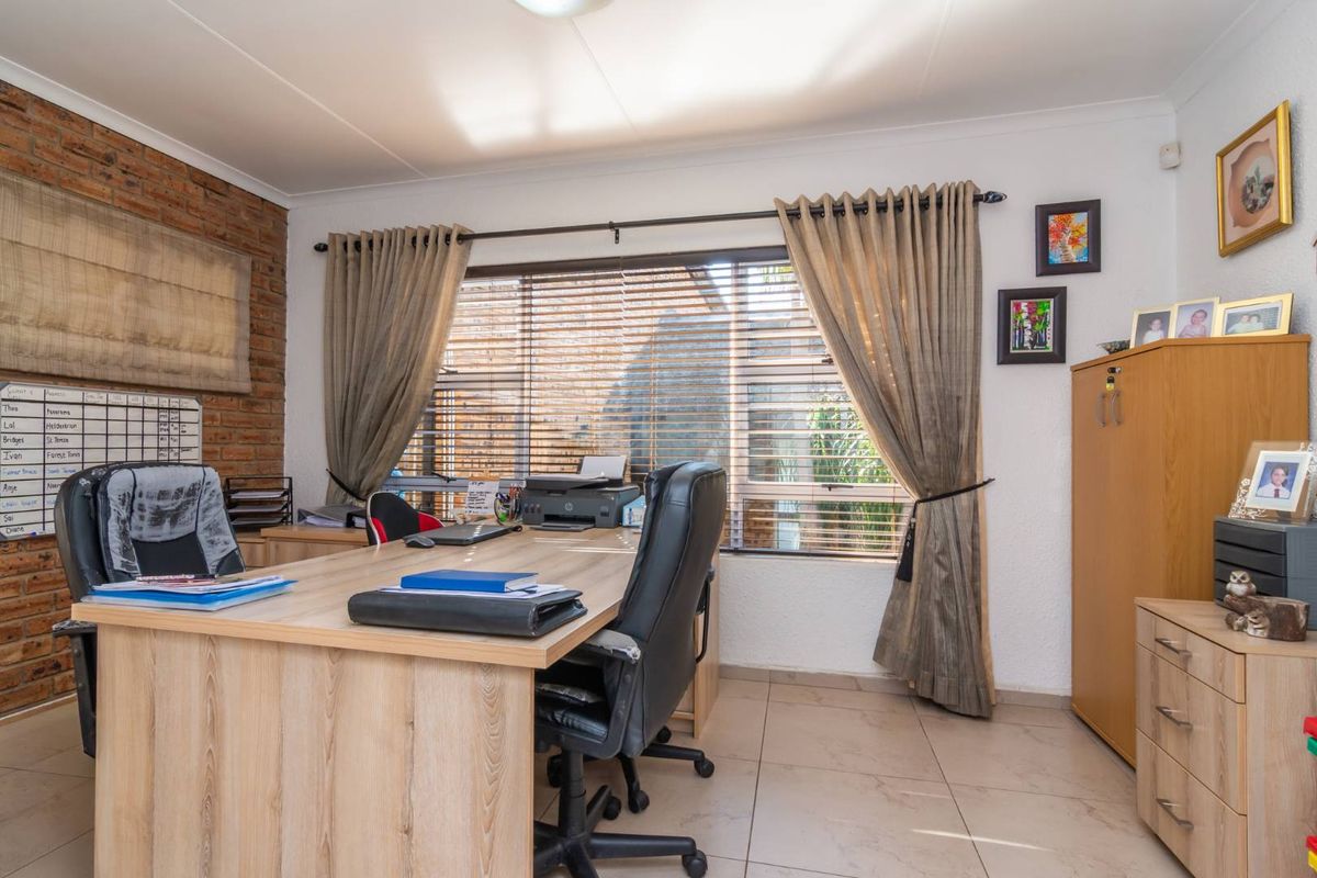 A functional home office featuring an exposed brick feature wall, tiled flooring, and large windows with venetian blinds for natural light control.