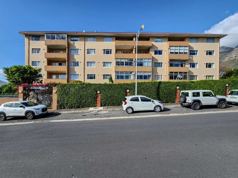 Front exterior view of Breda Park apartment complex, featuring a multi-story building with balconies and a secure gated entrance behind a landscaped hedge.