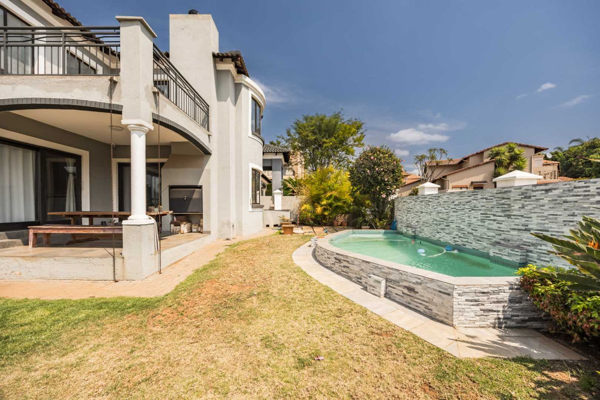 Rear exterior view featuring a covered patio with built-in braai, a raised stone-clad swimming pool, and a balcony overlooking the garden.