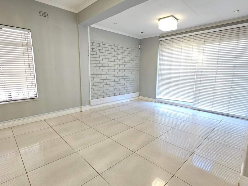 A bright, empty living room featuring large format floor tiles, an exposed brick feature wall, and sliding glass doors with venetian blinds.