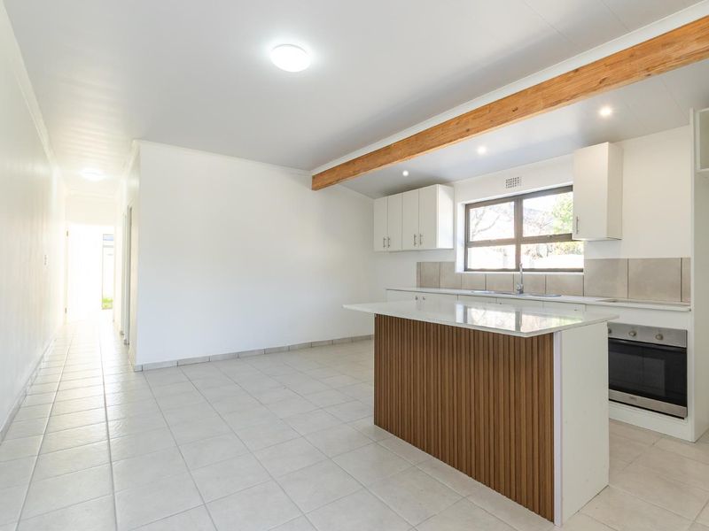A bright, open-plan kitchen area featuring a modern island, built-in oven, tiled flooring, and an exposed timber beam, offering a clean and functional space.