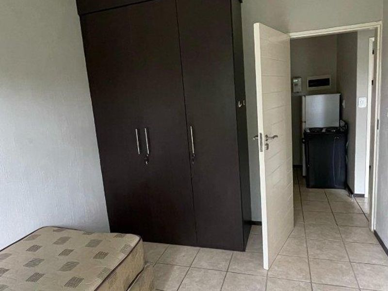A bedroom featuring tiled flooring and a dark wood built-in wardrobe, with an adjacent view into a small kitchen area.