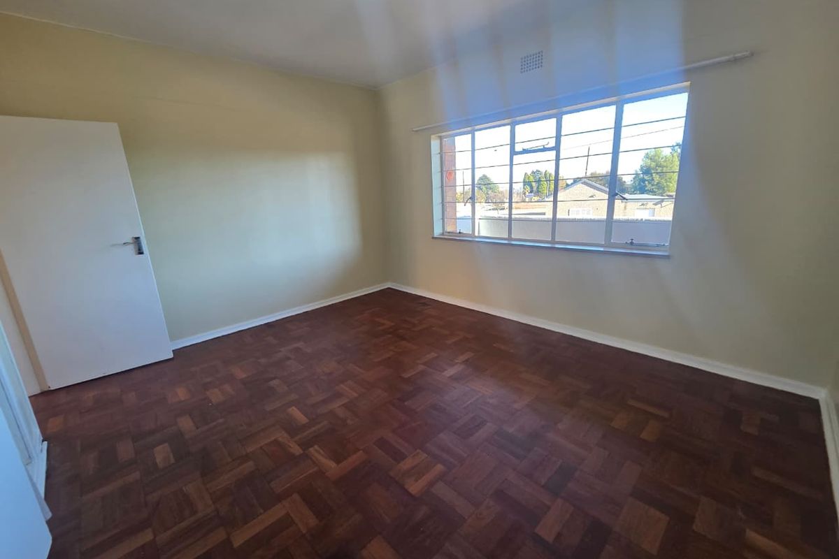 A bright, empty bedroom featuring classic parquet wood flooring, large windows with security bars, and neutral wall finishes.
