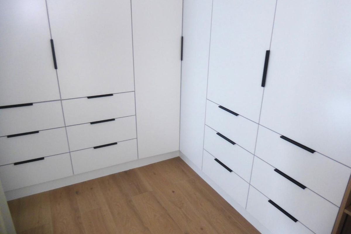 A clean and modern bedroom featuring extensive white built-in cupboards with matte black handles and light wood-look laminate flooring.