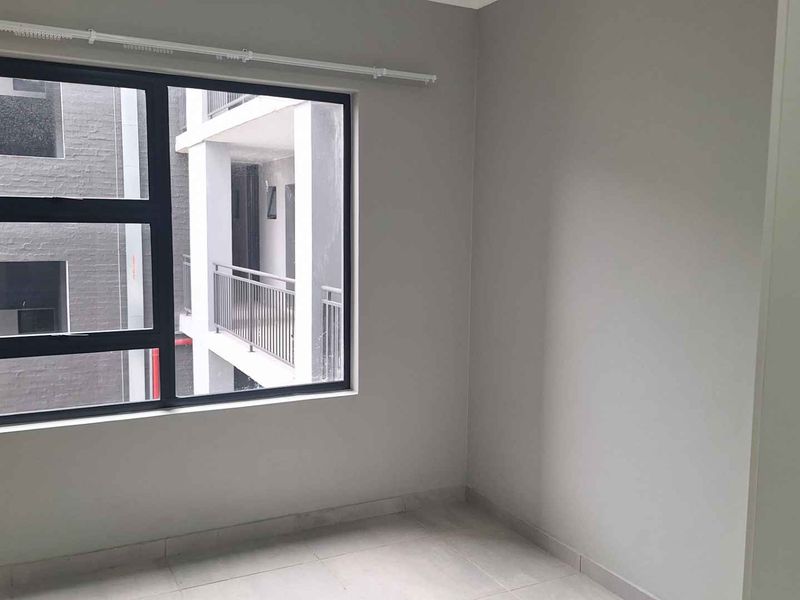 A bright, empty bedroom featuring neutral wall tones, tiled flooring, and a large aluminium-framed window overlooking the apartment complex.