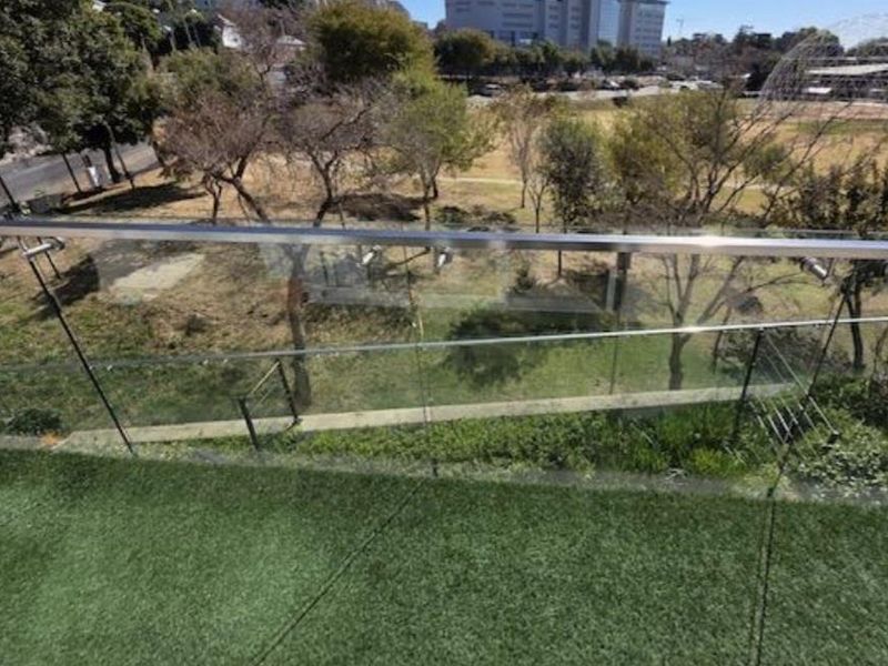A modern balcony featuring a sleek glass balustrade with a stainless steel handrail and artificial turf flooring, overlooking a green park area.