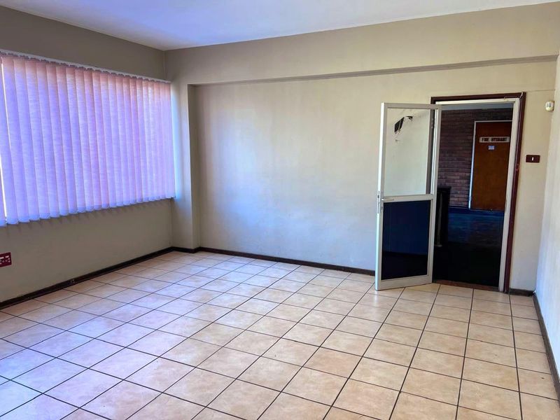 An empty office space featuring tiled flooring, vertical blinds, and an aluminium framed door leading to a hallway.