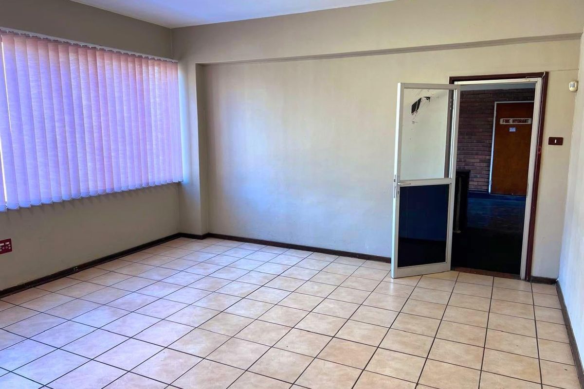 An empty office space featuring tiled flooring, vertical blinds, and an aluminium framed door leading to a hallway.