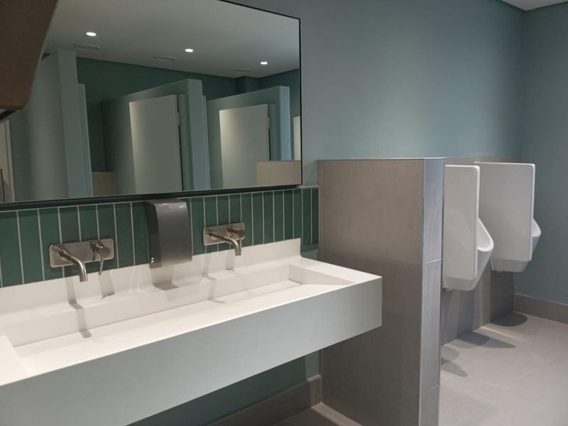 Modern commercial bathroom featuring a sleek wall-mounted trough sink, wall-mounted faucets, and clean tiled finishes with integrated urinals.