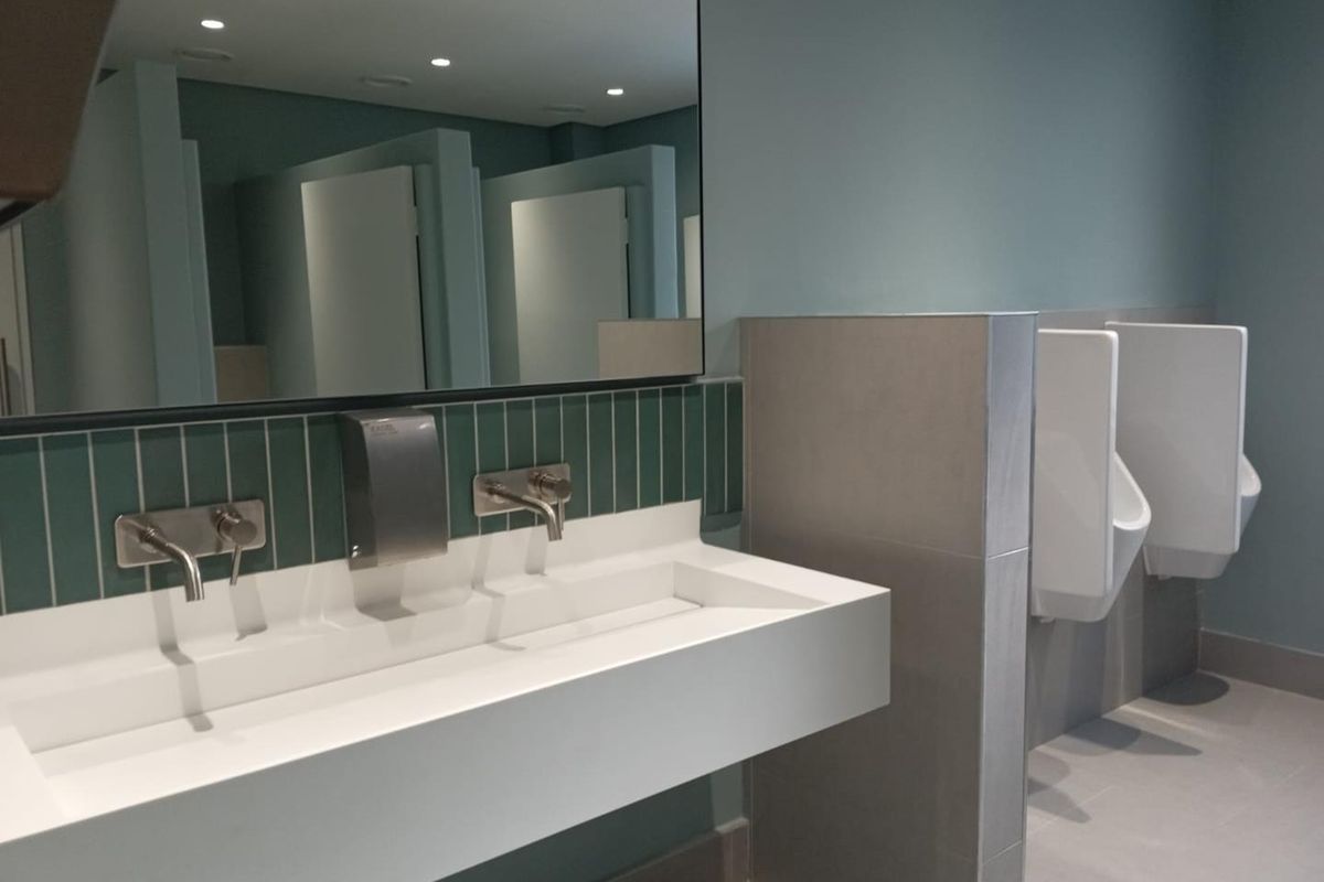 Modern commercial bathroom featuring a sleek wall-mounted trough sink, wall-mounted faucets, and clean tiled finishes with integrated urinals.