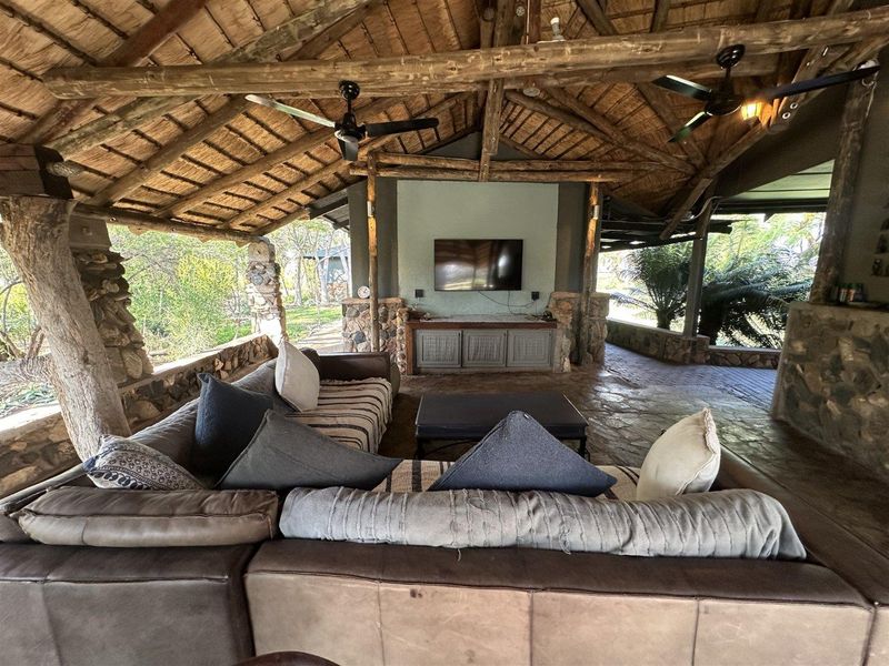 Spacious covered outdoor patio featuring a rustic thatched roof, exposed timber beams, stone masonry walls, and stone flooring, ideal for relaxed outdoor living.