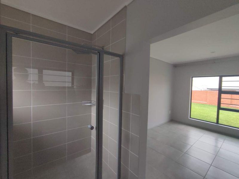 Modern bathroom featuring a tiled shower enclosure with a glass door, adjacent to a bright, tiled living area with large windows.