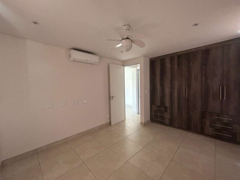 A spacious, empty bedroom featuring neutral tiled flooring, built-in wooden cupboards, a ceiling fan, and a wall-mounted air conditioning unit.