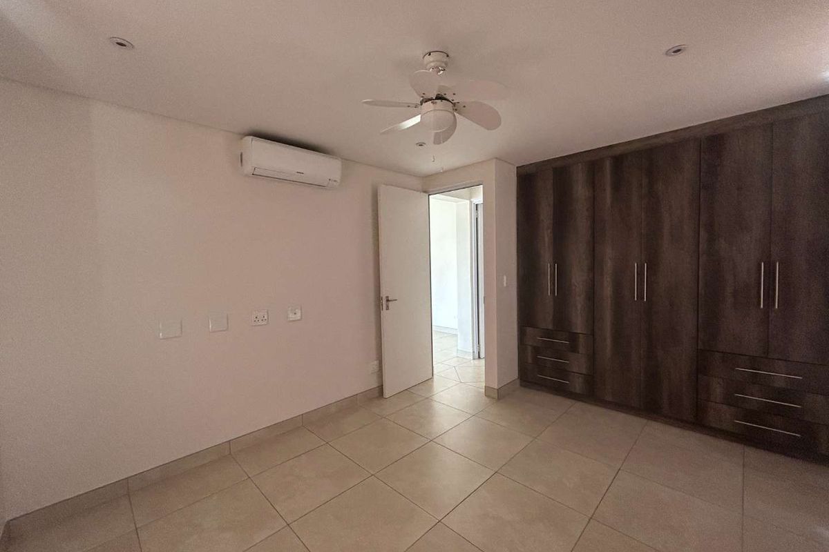 A spacious, empty bedroom featuring neutral tiled flooring, built-in wooden cupboards, a ceiling fan, and a wall-mounted air conditioning unit.