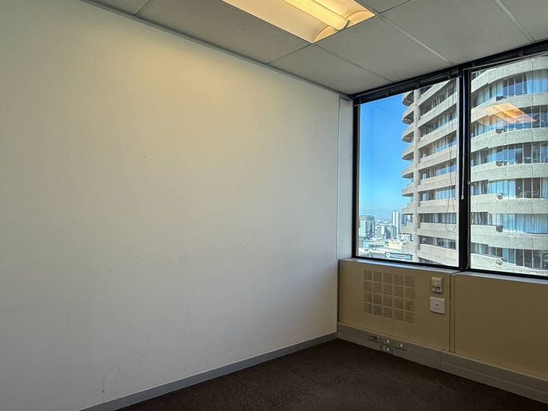 An empty office space featuring large windows with city views, suspended ceiling, and carpet flooring, suitable for a professional workspace.