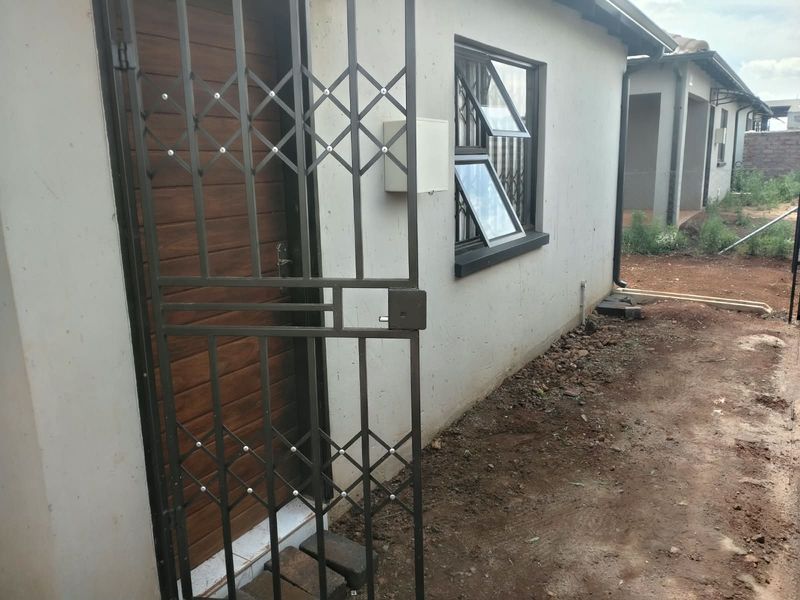 Rear exterior view of the property featuring a security gate, timber-look entrance door, and aluminium-framed windows, with unfinished garden area.