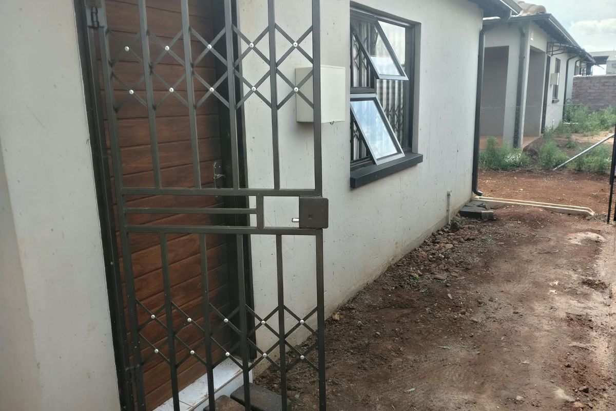 Rear exterior view of the property featuring a security gate, timber-look entrance door, and aluminium-framed windows, with unfinished garden area.