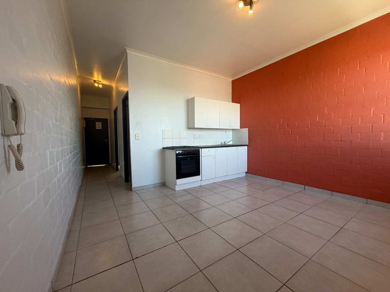 Open-plan living area featuring tiled flooring, an exposed brick feature wall, and a compact kitchenette with built-in cabinetry and appliances.