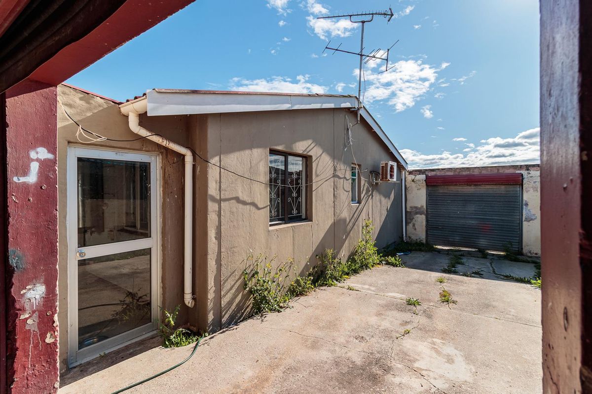 Rear exterior view showing a concrete patio area, security-fitted windows, a roller shutter garage door, and visible wear to the building's exterior finishes.